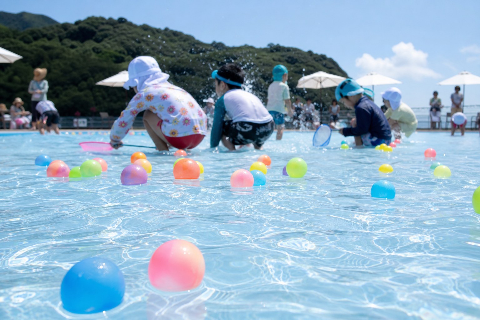 屋外プールの浅い子ども用プールで、水面に浮かぶカラフルなスーパーボールを子どもたちがすくい取っている様子
