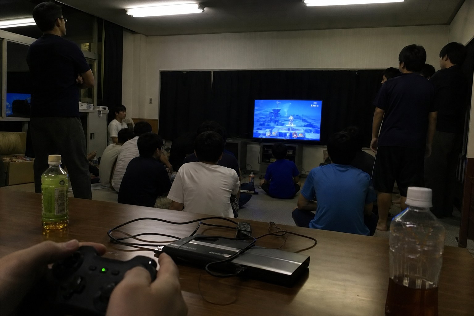 集会室のテレビの前に集まり、コントローラーを手に対戦ゲームを楽しむ子どもと大人たち
